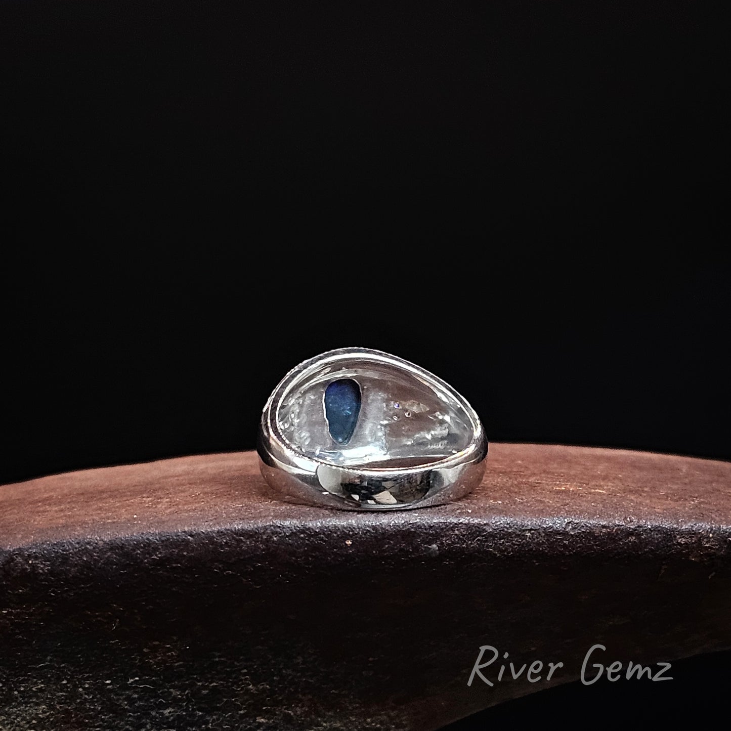 Rear view of a sterling silver ring showing the dark base tone of the inserted black opal, displayed on a dark wooden surface against a black background. The brand name 'River Gemz' is visible in the bottom right corner.