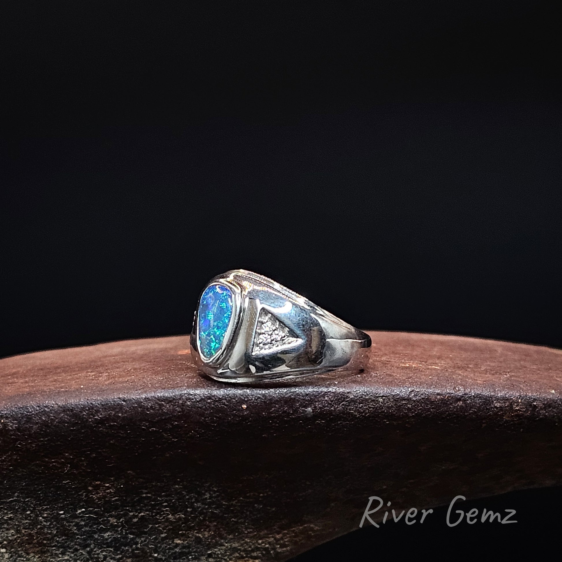 A sterling silver ring featuring a blue opal gemstone and a triangular inset with 3 white diamonds, displayed on a dark wooden surface against a black background.