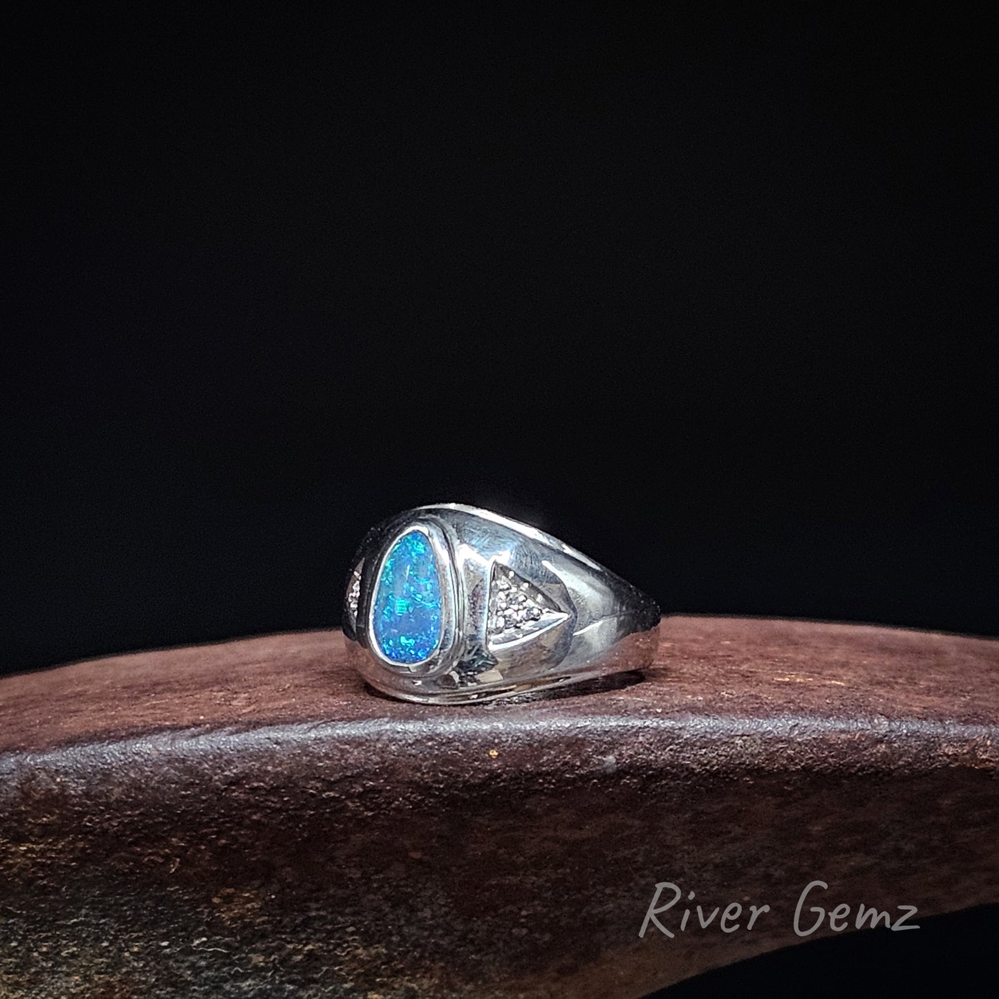 A sterling silver signet ring featuring a vibrant blue teardrop-shaped opal and a small triangle of 3 diamonds on the band, displayed on a dark brown wooden block against a black background.