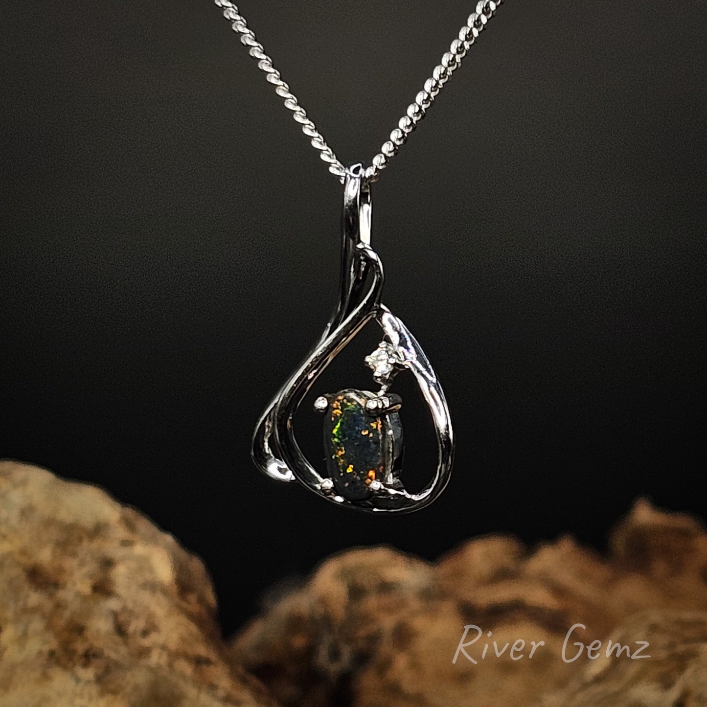 Silver pendant with a central gemstone on a dark background, featuring 'River Gemz' branding.