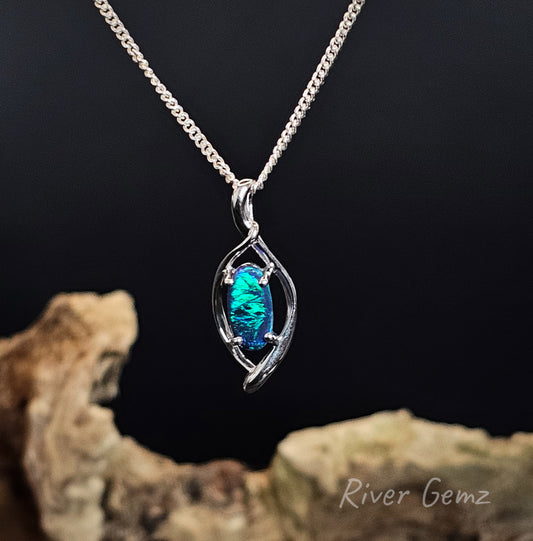 Green and blue oval shaped opal claw set in silver pendant with hidden bail against a black background and with a piece of drift wood at the base of the photograph including 'River Gemz' watermark.