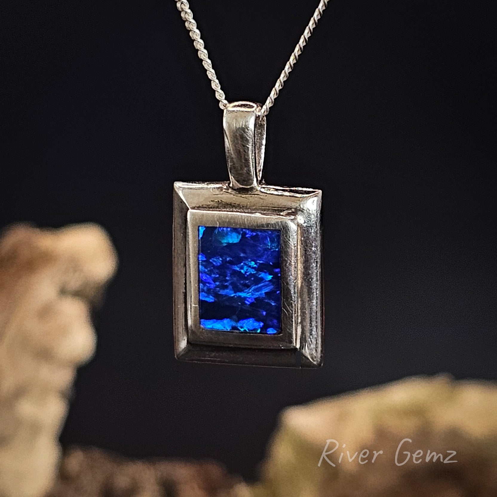 Silver pendant with blue stone on a dark background, featuring 'River Gemz' branding.