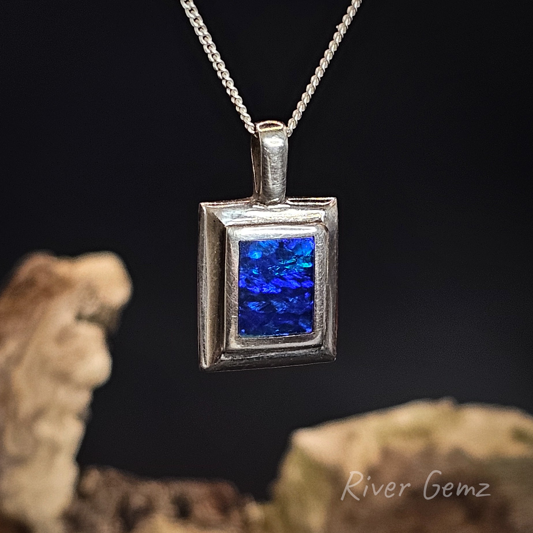 Silver pendant with blue stone on a chain against a dark background, featuring 'River Gemz' branding.