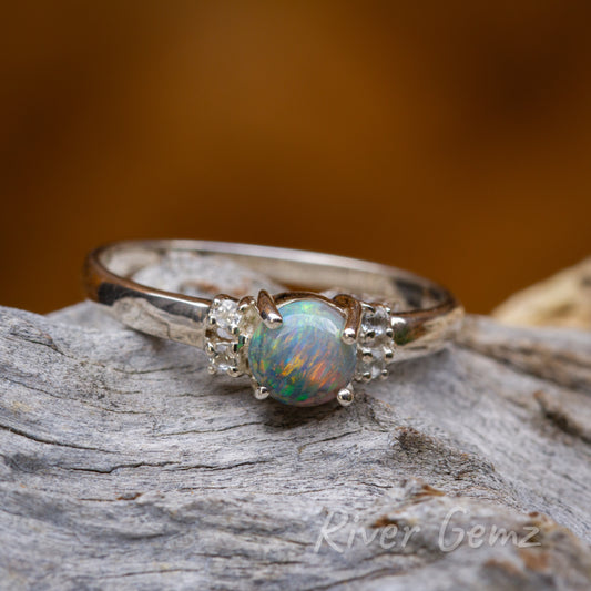 Round colourful black fire opal and 4 white topaz stones are held securely with four claws in the silver ring. The ring is lying on a weathered piece of wood.