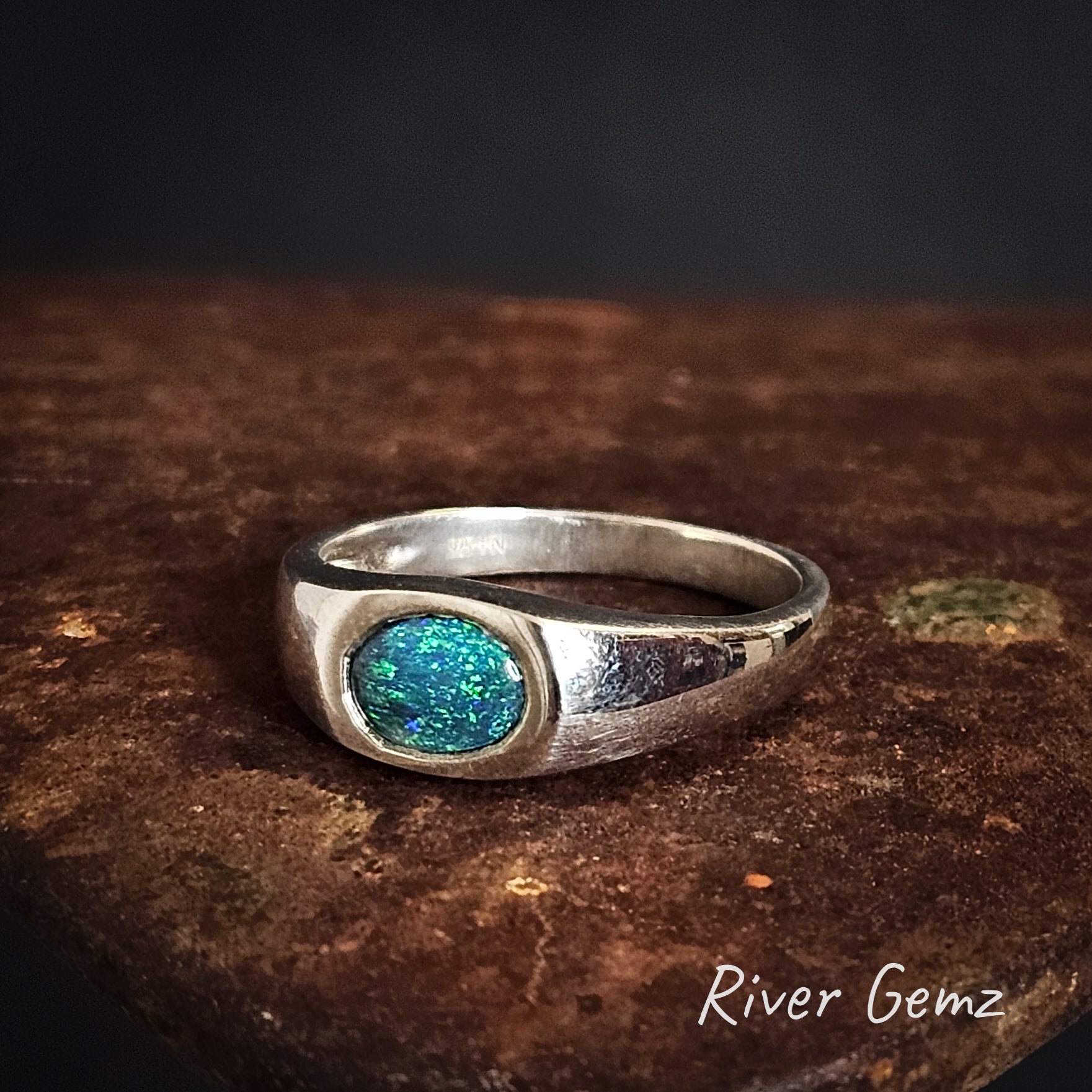 Silver ring with a blue black opal on a dark surface, featuring 'River Gemz' branding.
