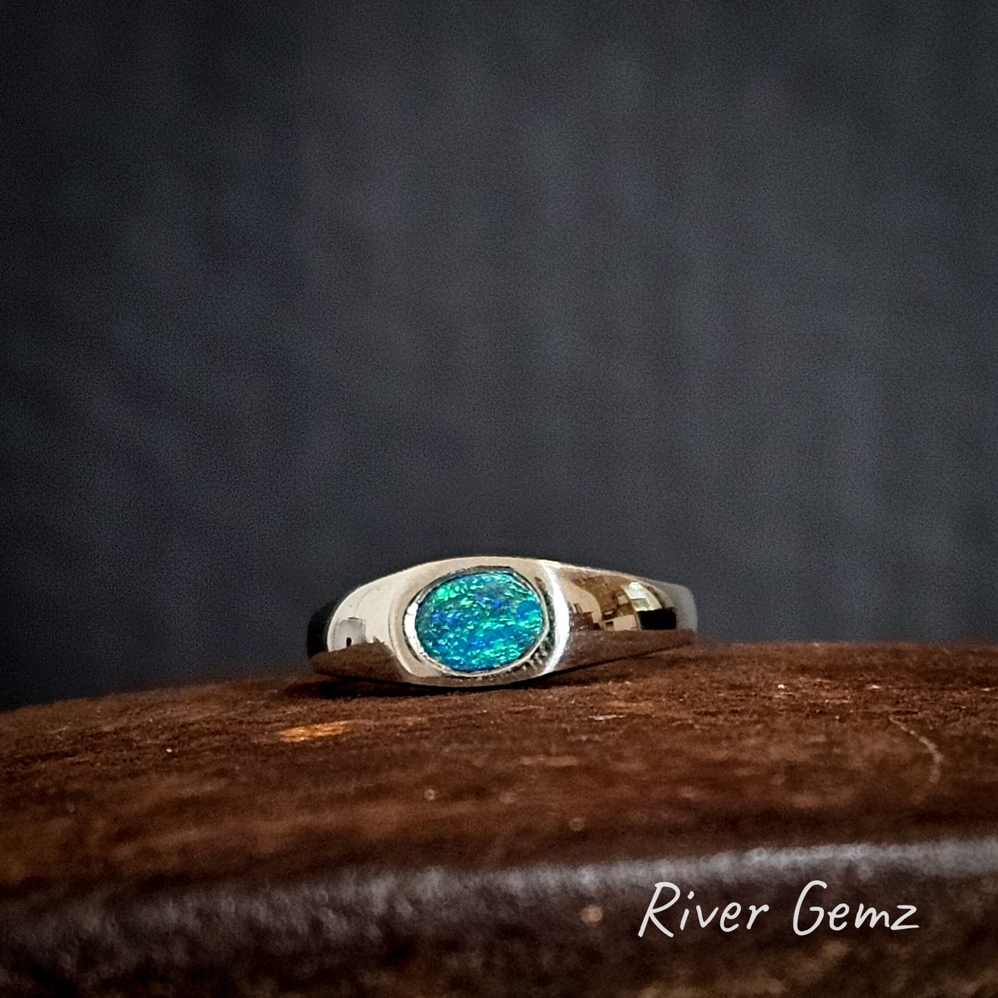 Silver ring with an oval shaped blue and green opal on a rust coloured surface, branded 'River Gemz'.