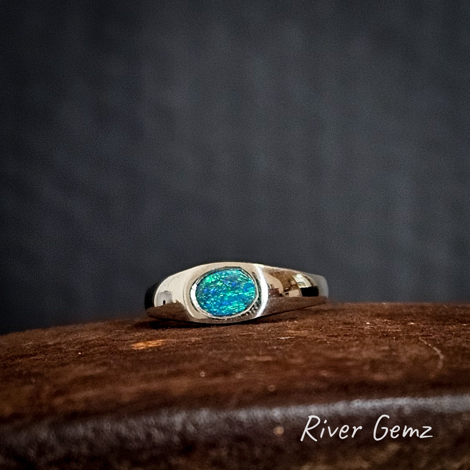 Silver ring with an oval shaped blue and green opal on a rust coloured surface, branded 'River Gemz'.