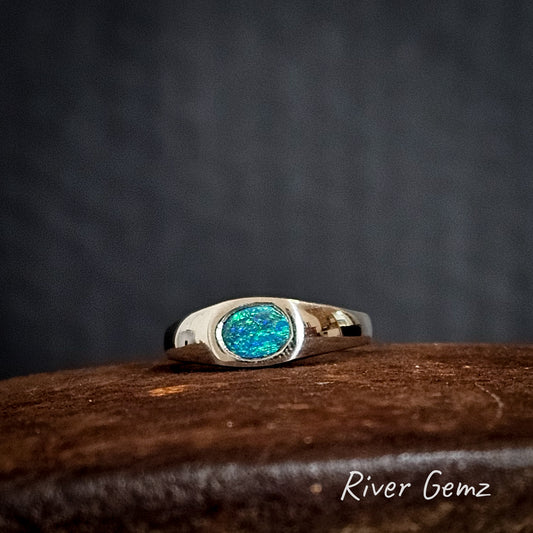 Silver ring with an oval shaped blue and green opal on a rust coloured surface, branded 'River Gemz'.