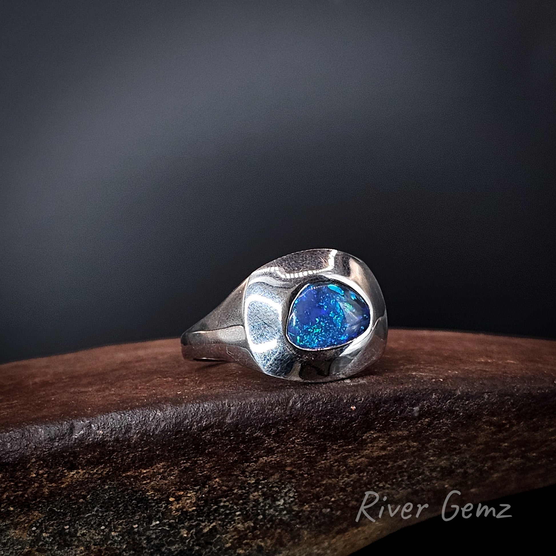 Silver ring with dark blue black opal on an rusty brown base with 'River Gemz' branding and black background.