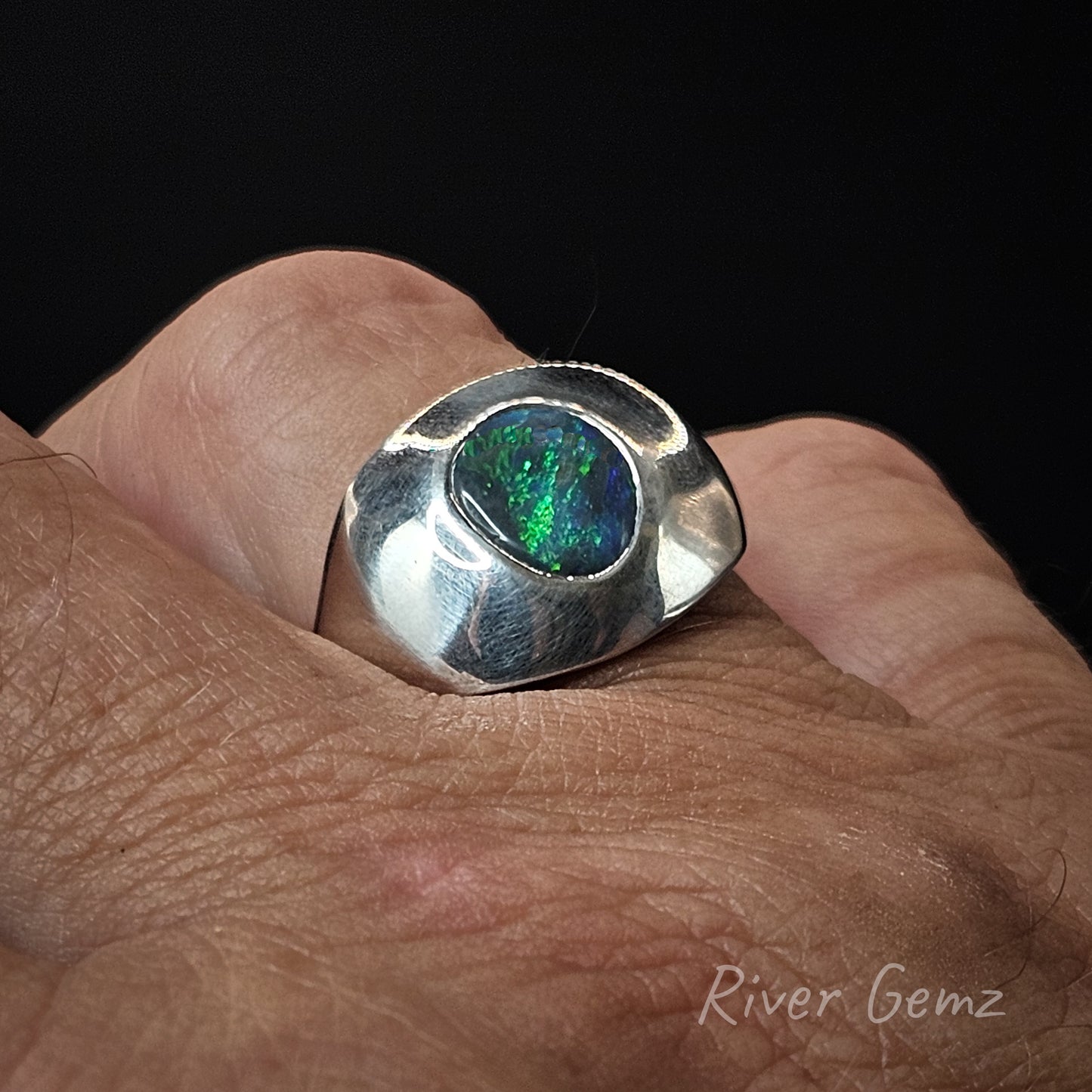 Silver ring with an opal stone on a hand, branded 'River Gemz'.
