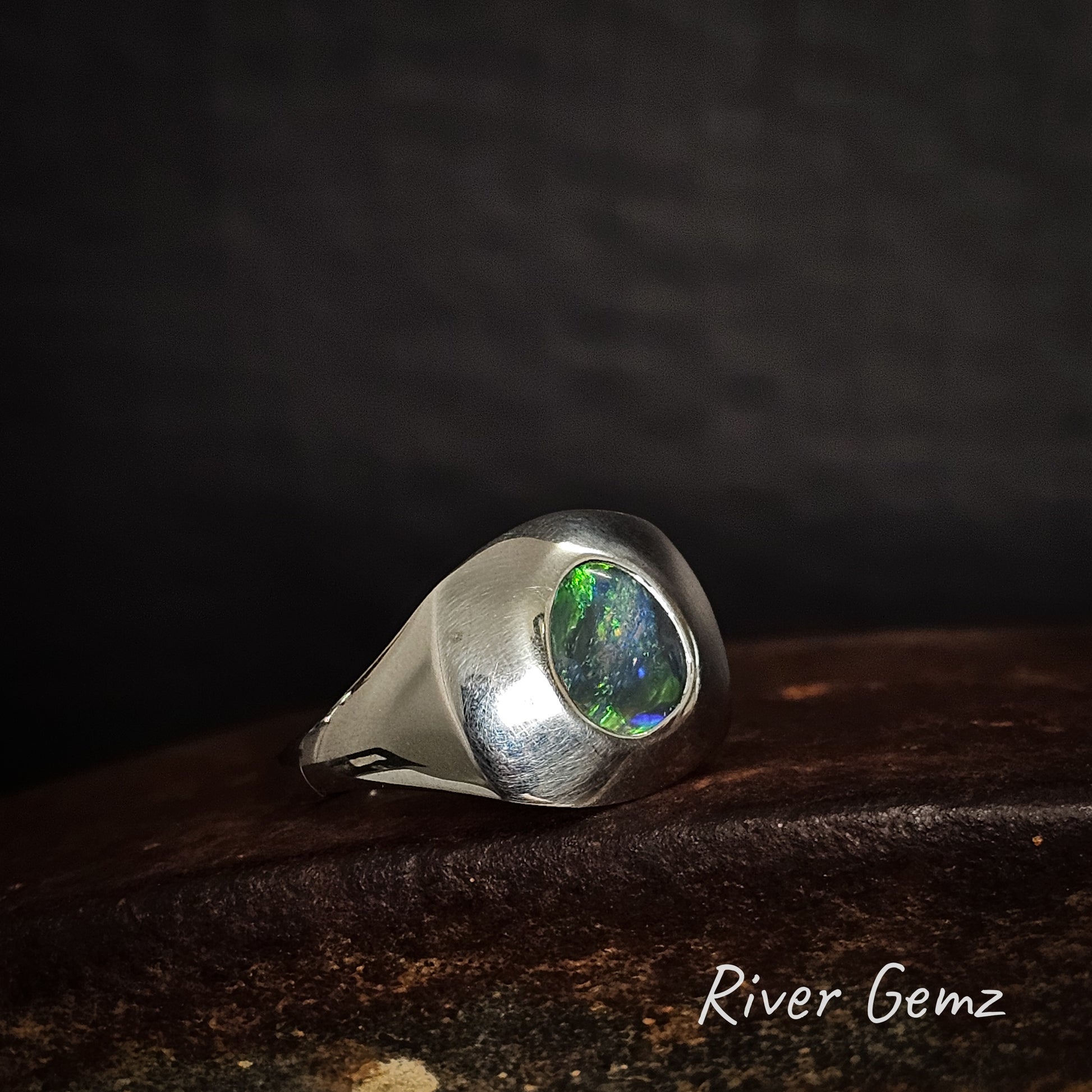 Men's silver ring with a green gemstone on a dark surface, branded 'River Gemz'.