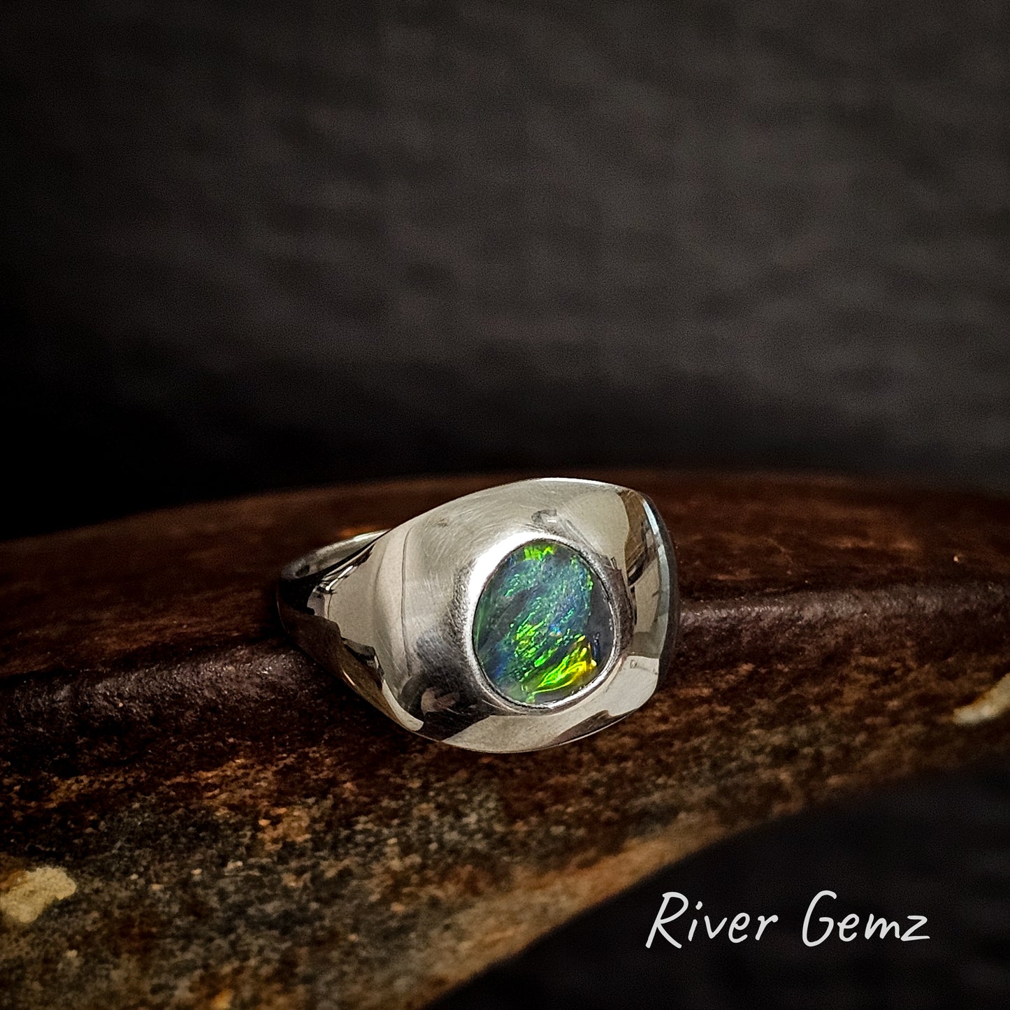 Silver ring with a green, yellow and blue dark opal on a dark background, featuring the brand 'River Gemz'.