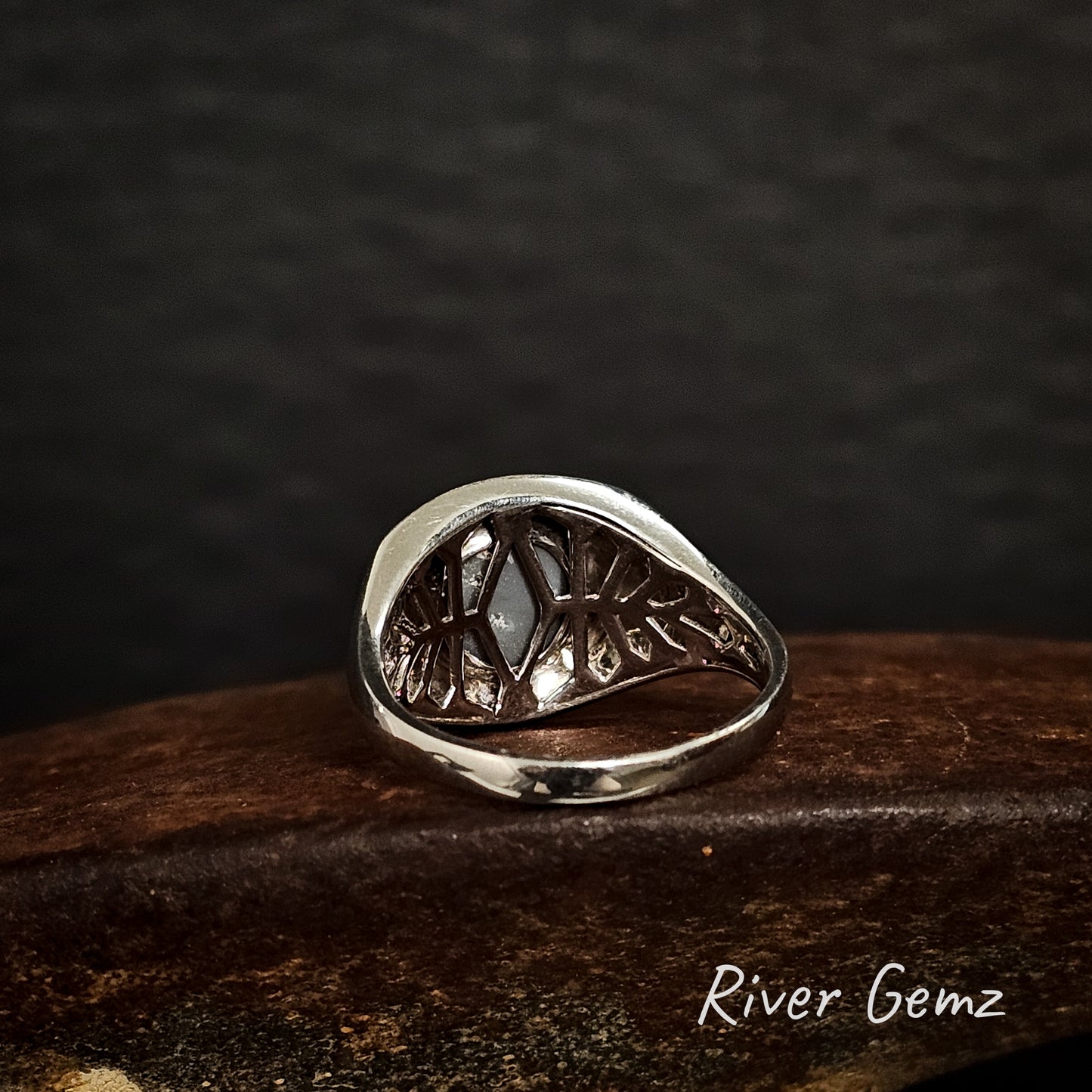 Back of silver ring shows the lattice work design used behind the black opal on a dark surface, branded 'River Gemz'.