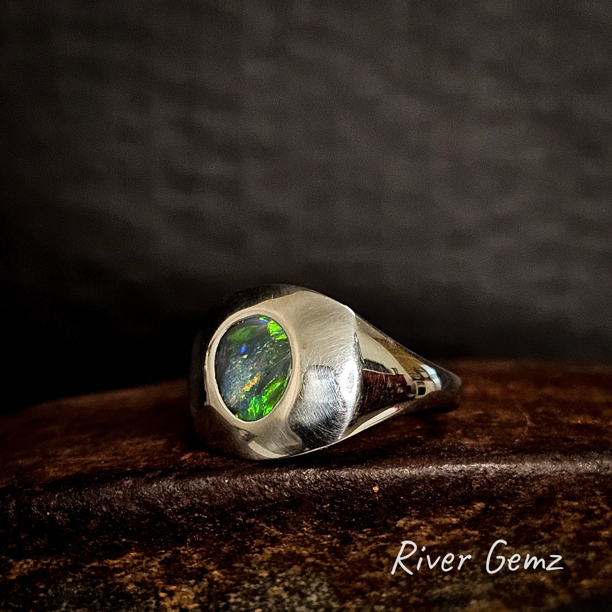 Silver ring with a green gemstone on a dark wooden surface, featuring 'River Gemz' branding.