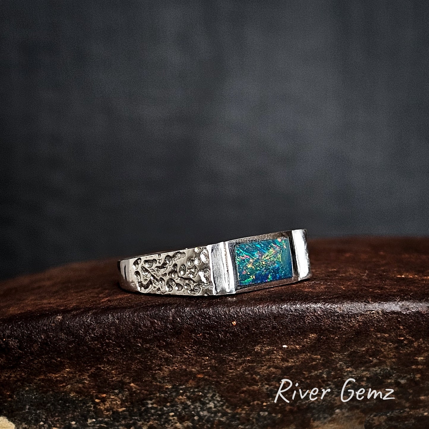 Silver ring with rectangular colourful black opal on a dark surface, branded 'River Gemz'.