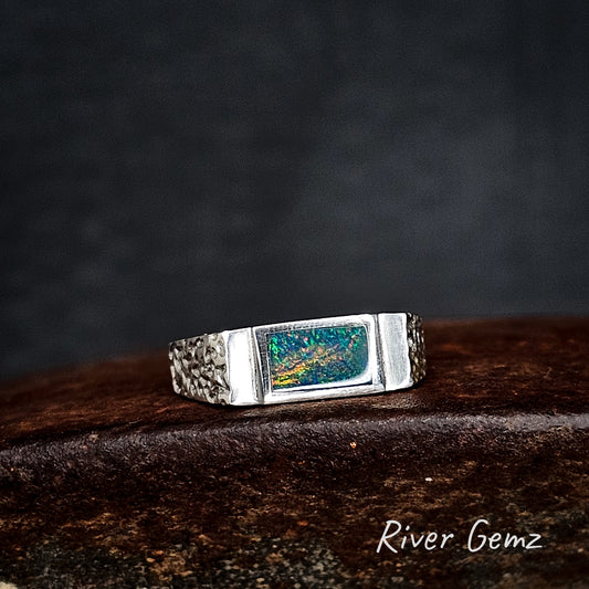 Silver ring with a rectangular multi-coloured black opal on a dark surface, branded 'River Gemz'.