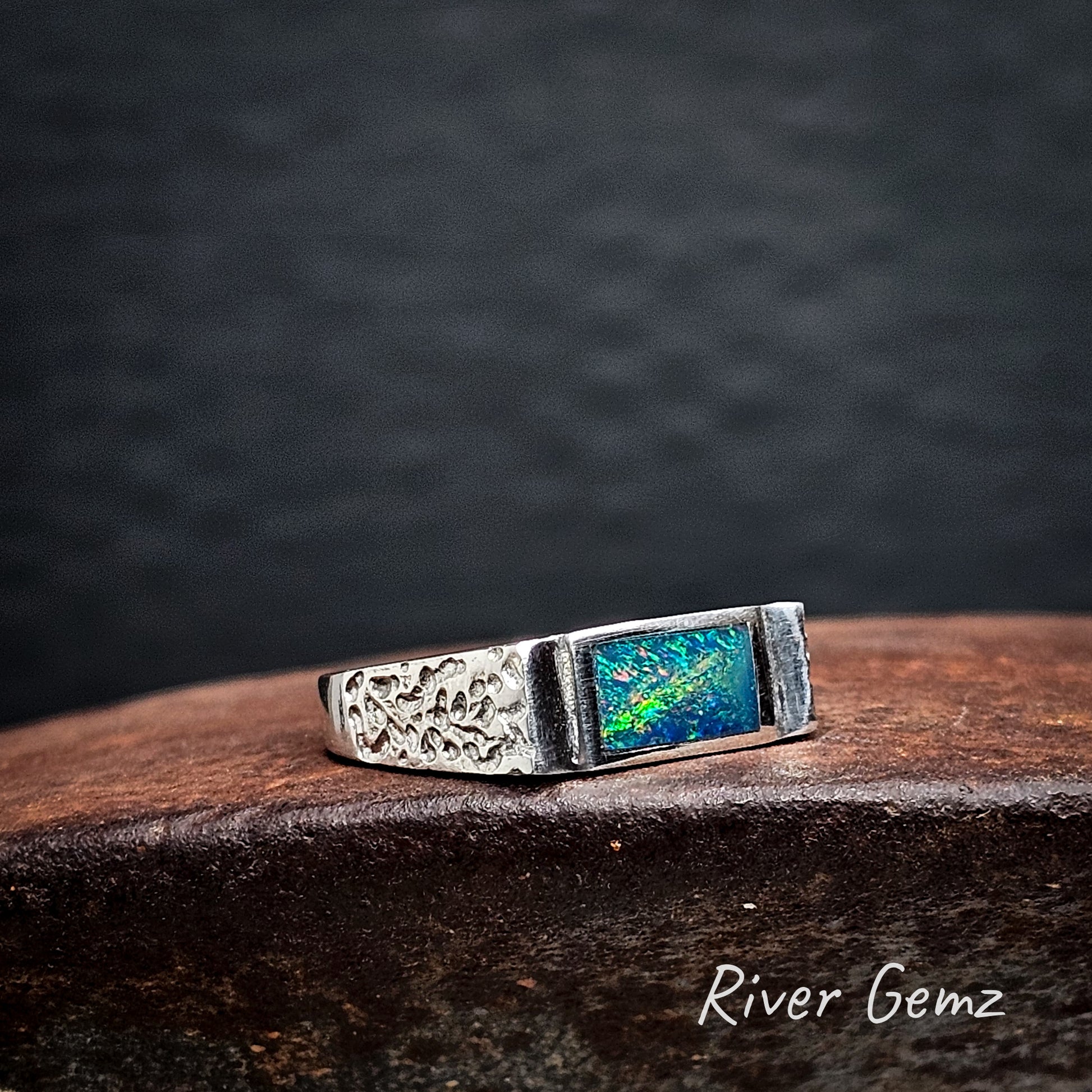 Silver ring with a colorful dark opal on a rusty brown surface, branded 'River Gemz'.