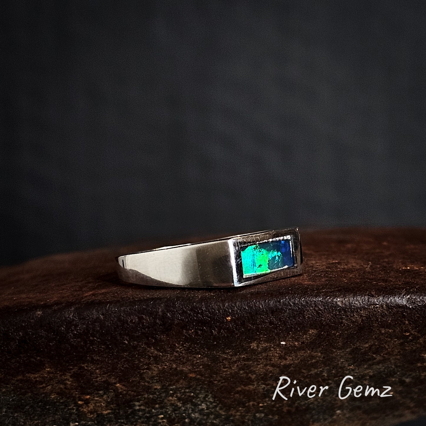 Silver ring with a rectangular shaped black opal on a dark background, branded 'River Gemz'.
