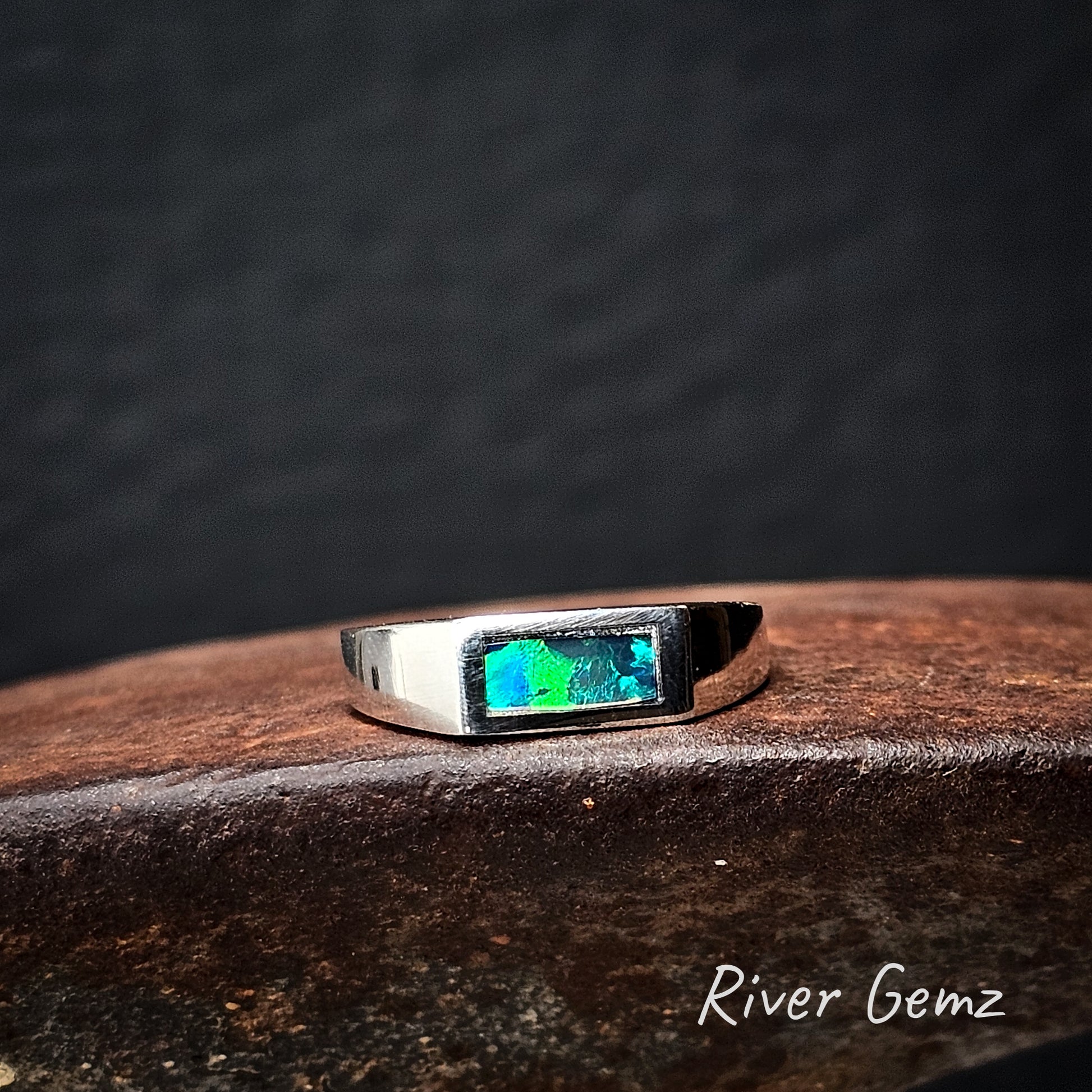 Silver ring with a green dark opal on a cast iron surface, branded 'River Gemz'.