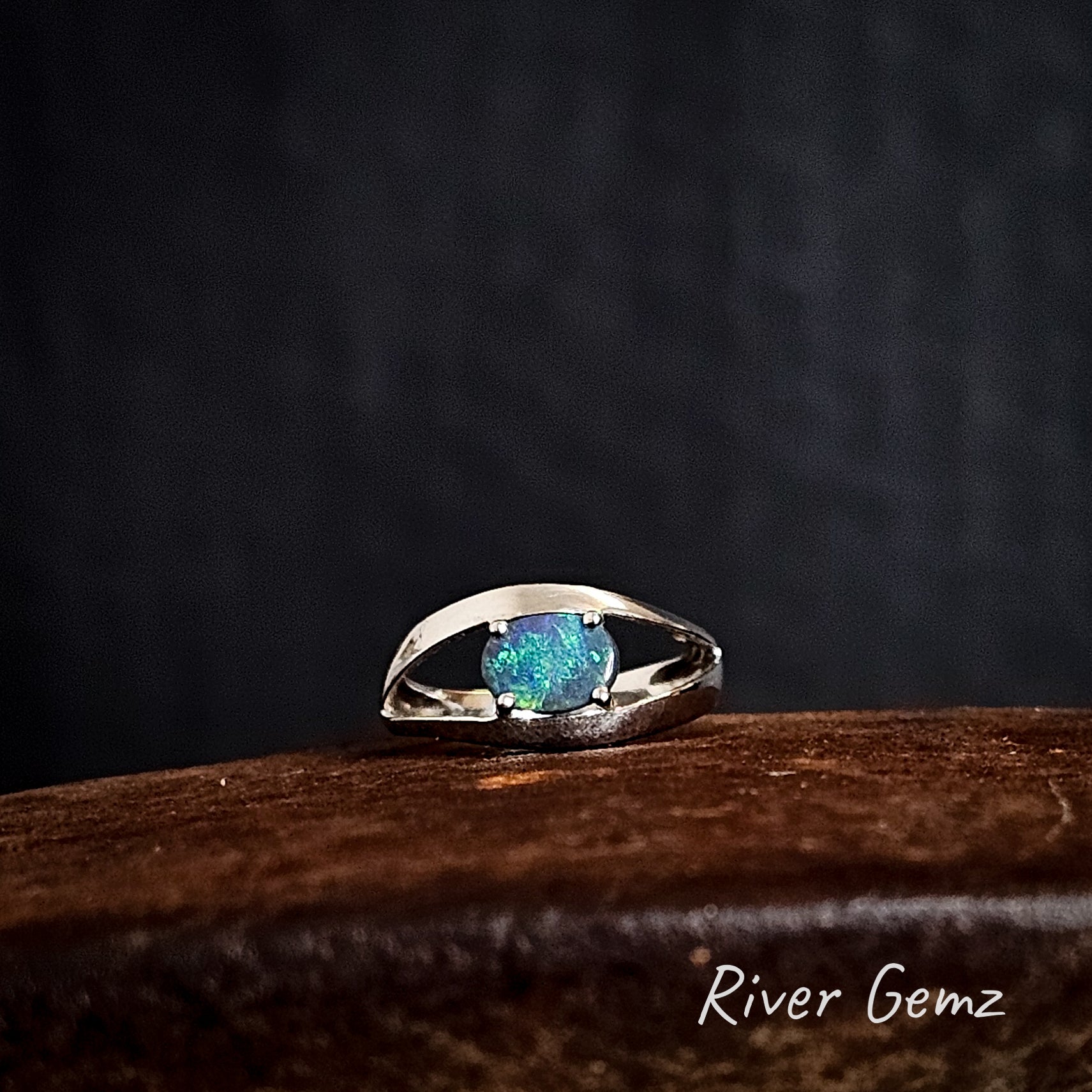 Oval shaped dark opal claw set in a silver ring on a rusty coloured surface with "River Gemz" branding.