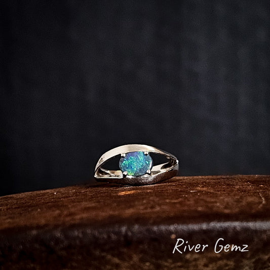 Oval shaped dark opal claw set in a silver ring on a rusty coloured surface with "River Gemz" branding.