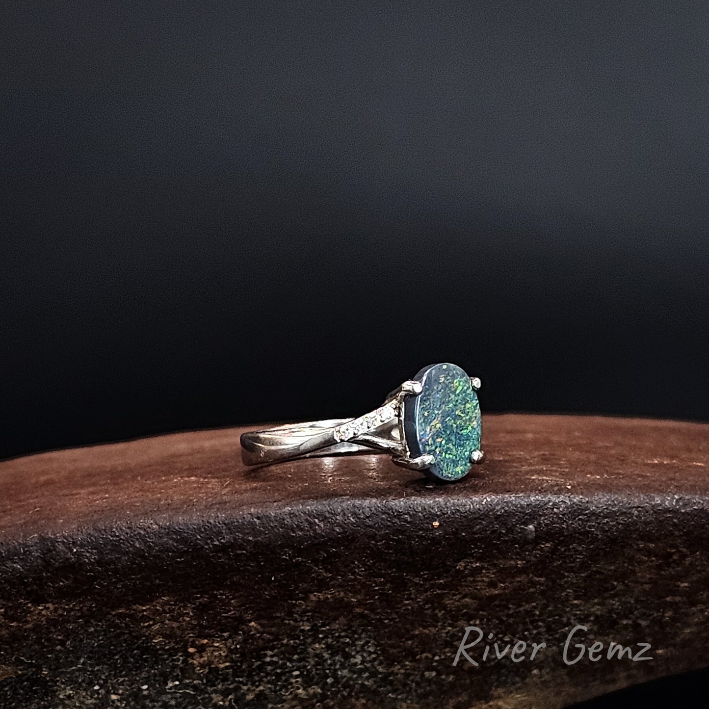 Oval shaped dark opal claw set in silver ring on a rusty shoe anvil with 'River Gemz' branding.
