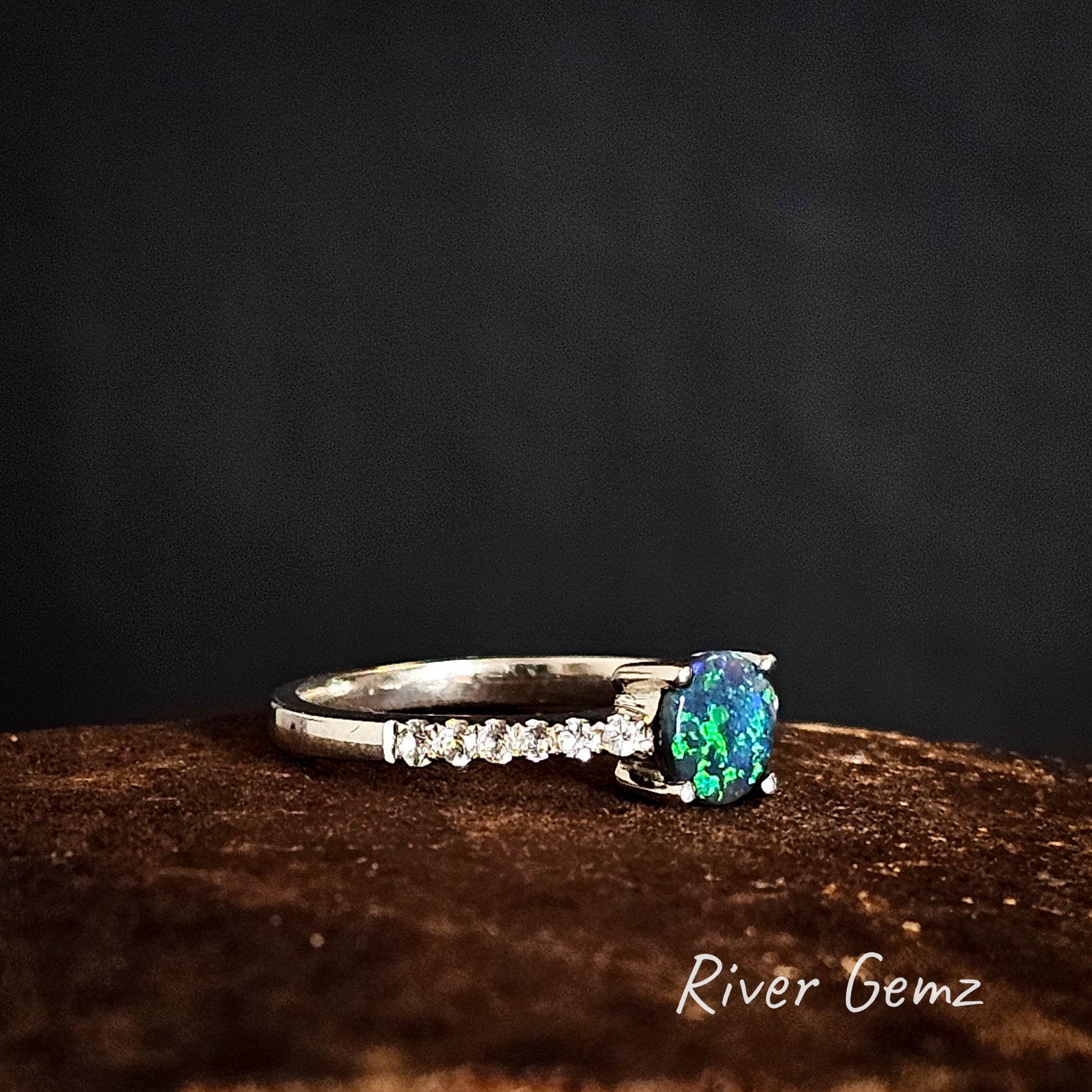 Silver ring with a green dark opal and  white topaz stones on the shoulders on a dark background, branded 'River Gemz'.