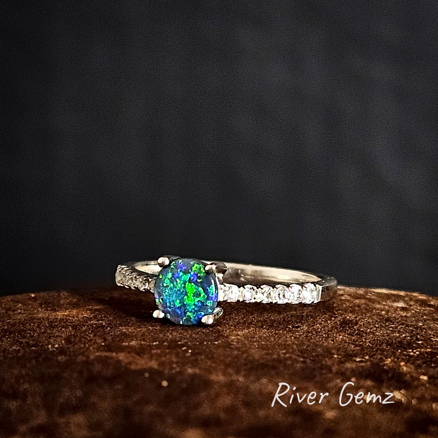 Ring with a blue and green round 5 mm gemstone on a dark background, branded 'River Gemz'.