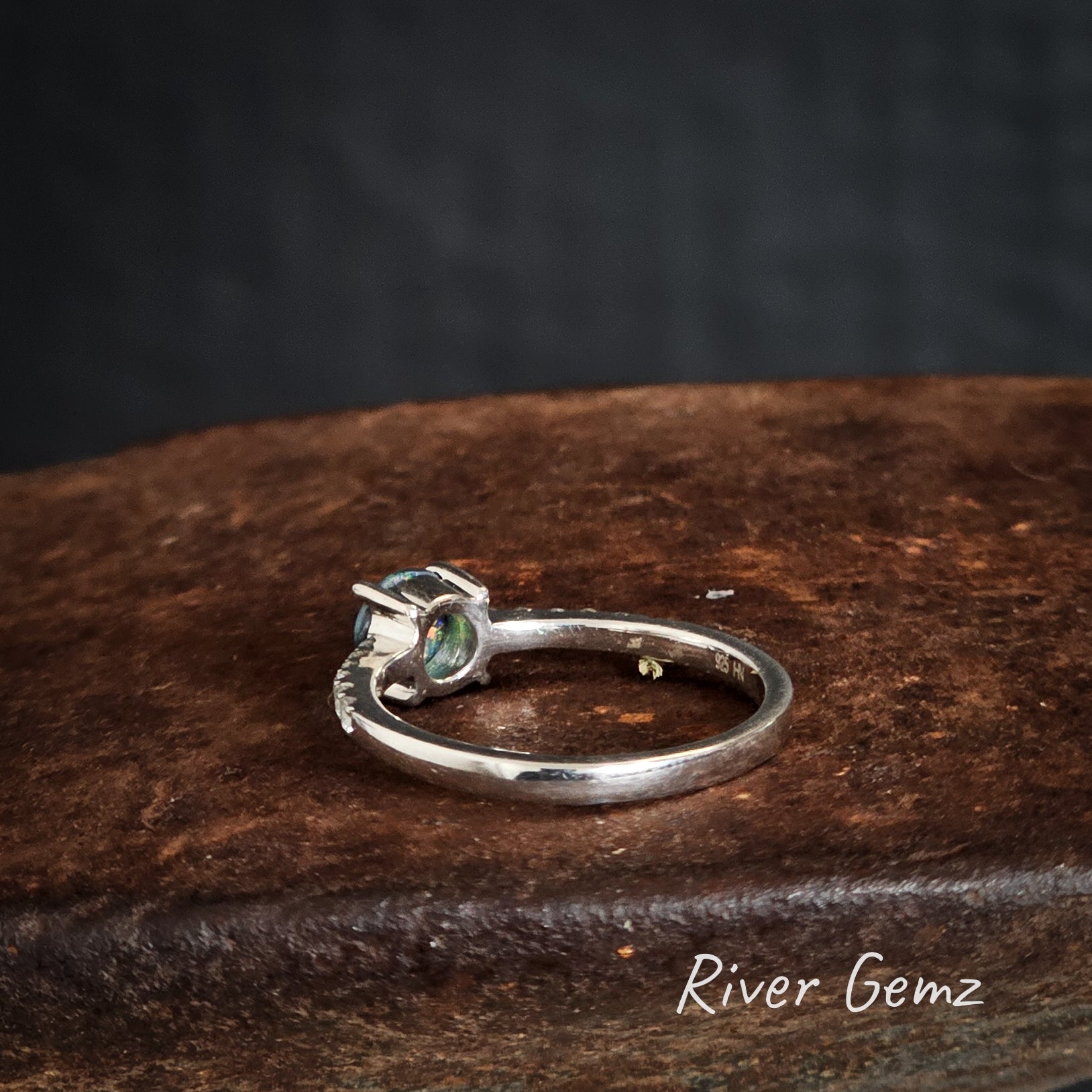 Back of silver ring with a gemstone on a rusty shoe anvil, branded 'River Gemz'.