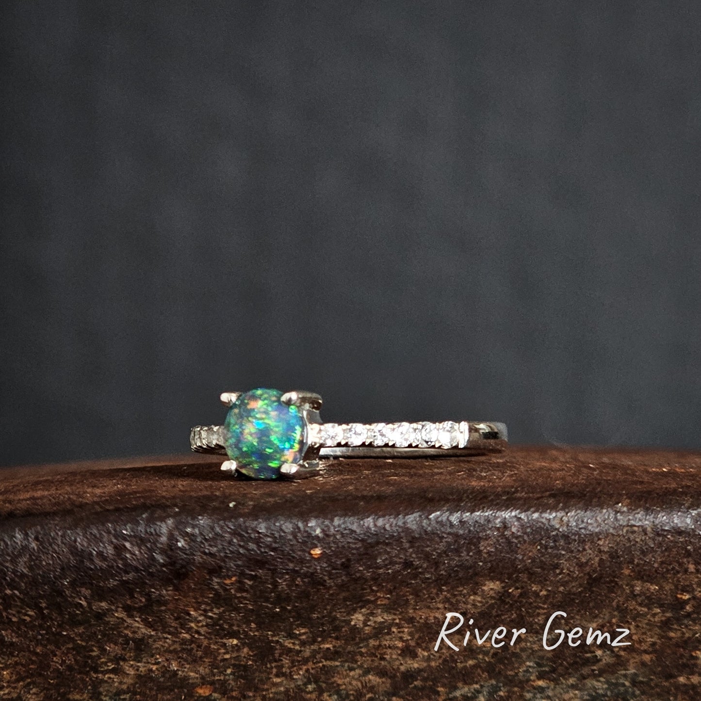 Silver ring with a multi-coloured black opal on a dark surface, branded 'River Gemz'.