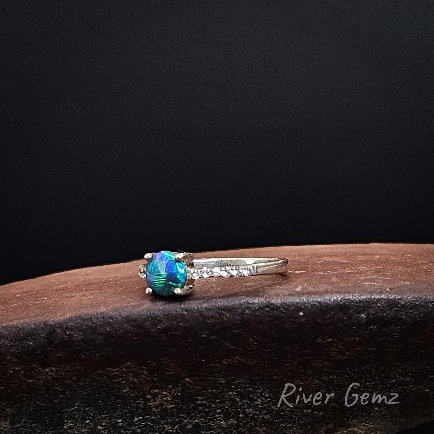 Blue and green black opal claw set in silver ring on a rusty shoe anvil with 'River Gemz' branding.