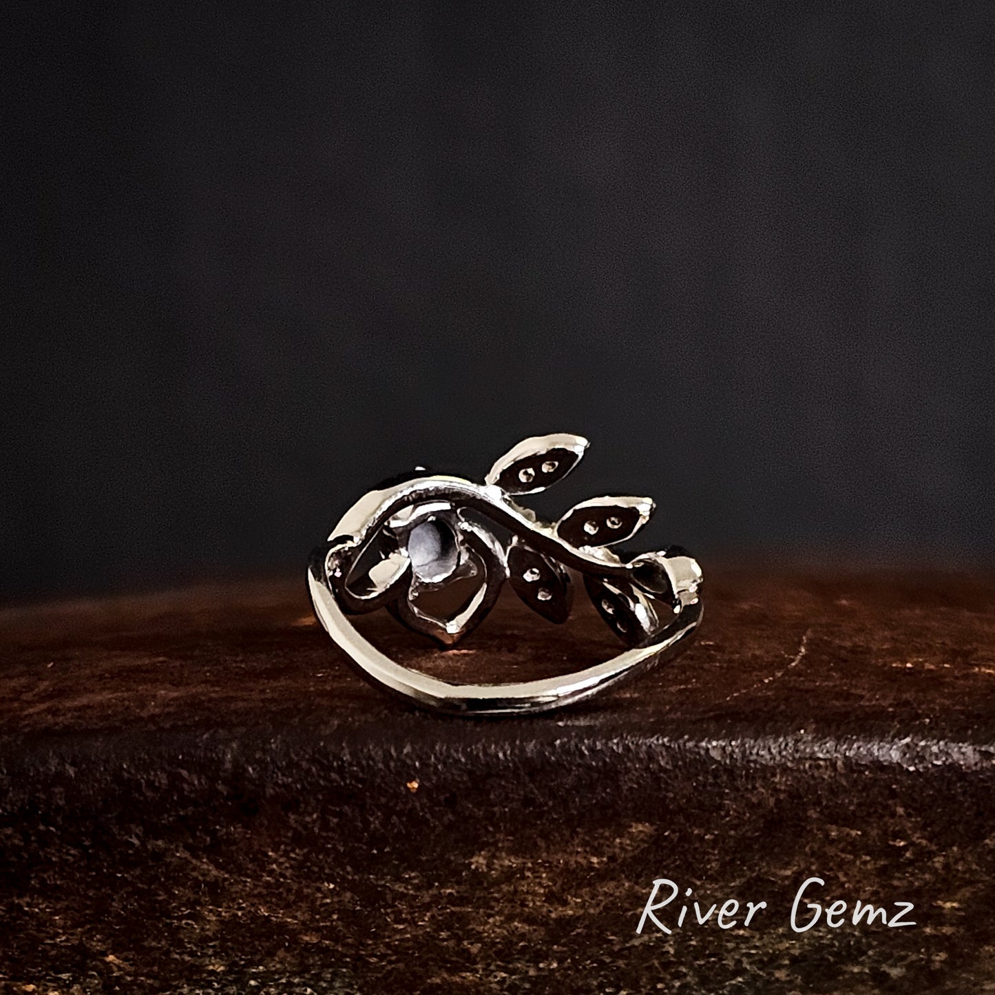 Back of silver ring with leaf design showing the dark tone of the included black opal on a rusty surface, branded 'River Gemz'.