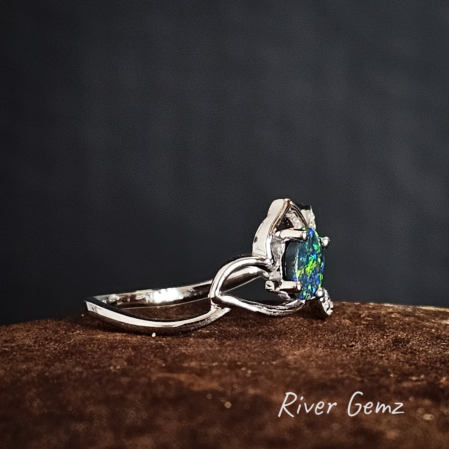 Silver ring with a blue & green black opal on a dark background, branded 'River Gemz'.