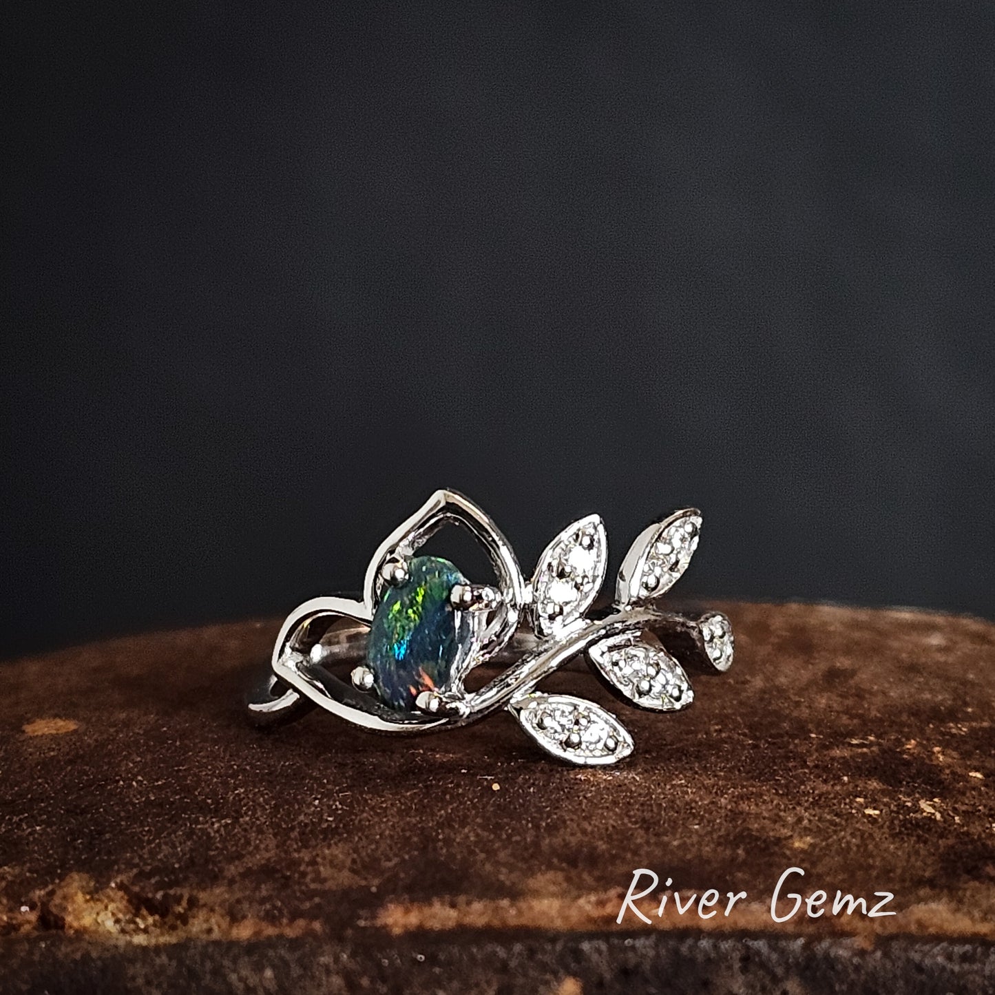 Silver ring with a blue/green dark opal with a splash of orange on a dark background, branded 'River Gemz'.