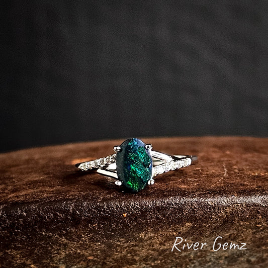 Ring with a dark green black opal and white topaz shoulders on a dark brown surface, branded 'River Gemz'.