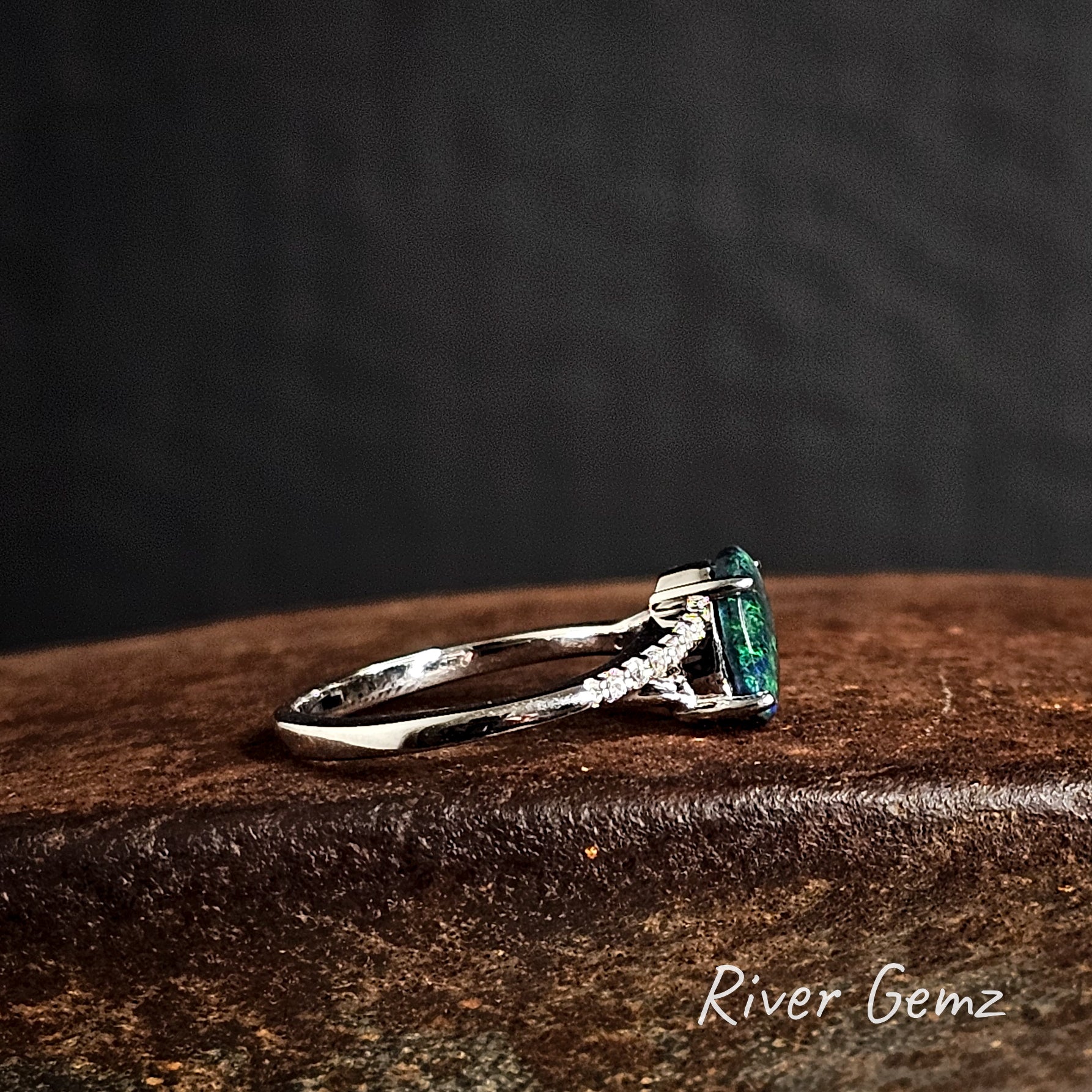 Side view of silver ring with a green solid black opal on a dark surface, featuring 'River Gemz' branding.