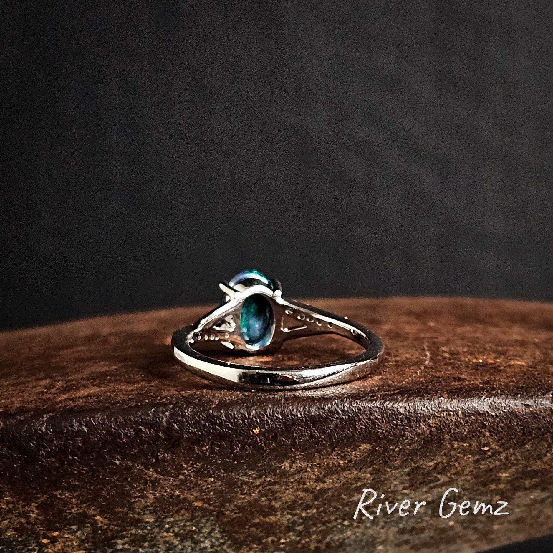 Rear view of the silver ring shows the dark tone of the included black opal on a cast iron surface, branded 'River Gemz'.