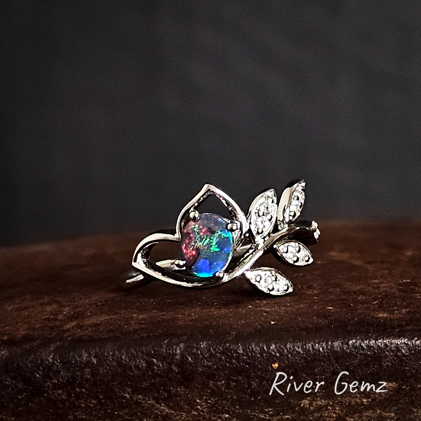 Silver ring with a colorful gemstone on a dark background, branded 'River Gemz'.