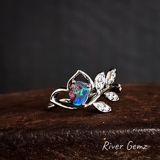 Silver ring with a colorful gemstone on a dark background, branded 'River Gemz'.