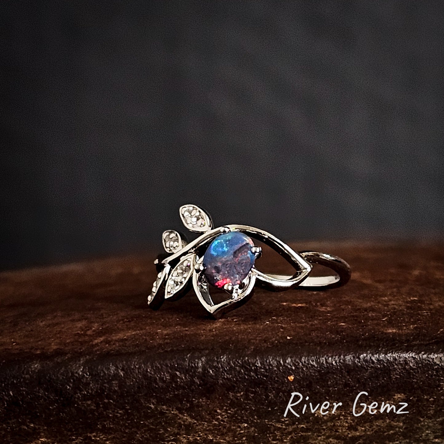 Silver ring with a colourful dark opal on a dark rust coloured surface, branded 'River Gemz'.