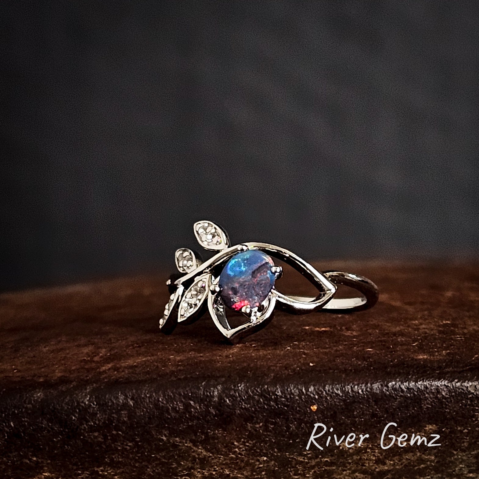 Silver ring with a colourful dark opal on a dark rust coloured surface, branded 'River Gemz'.