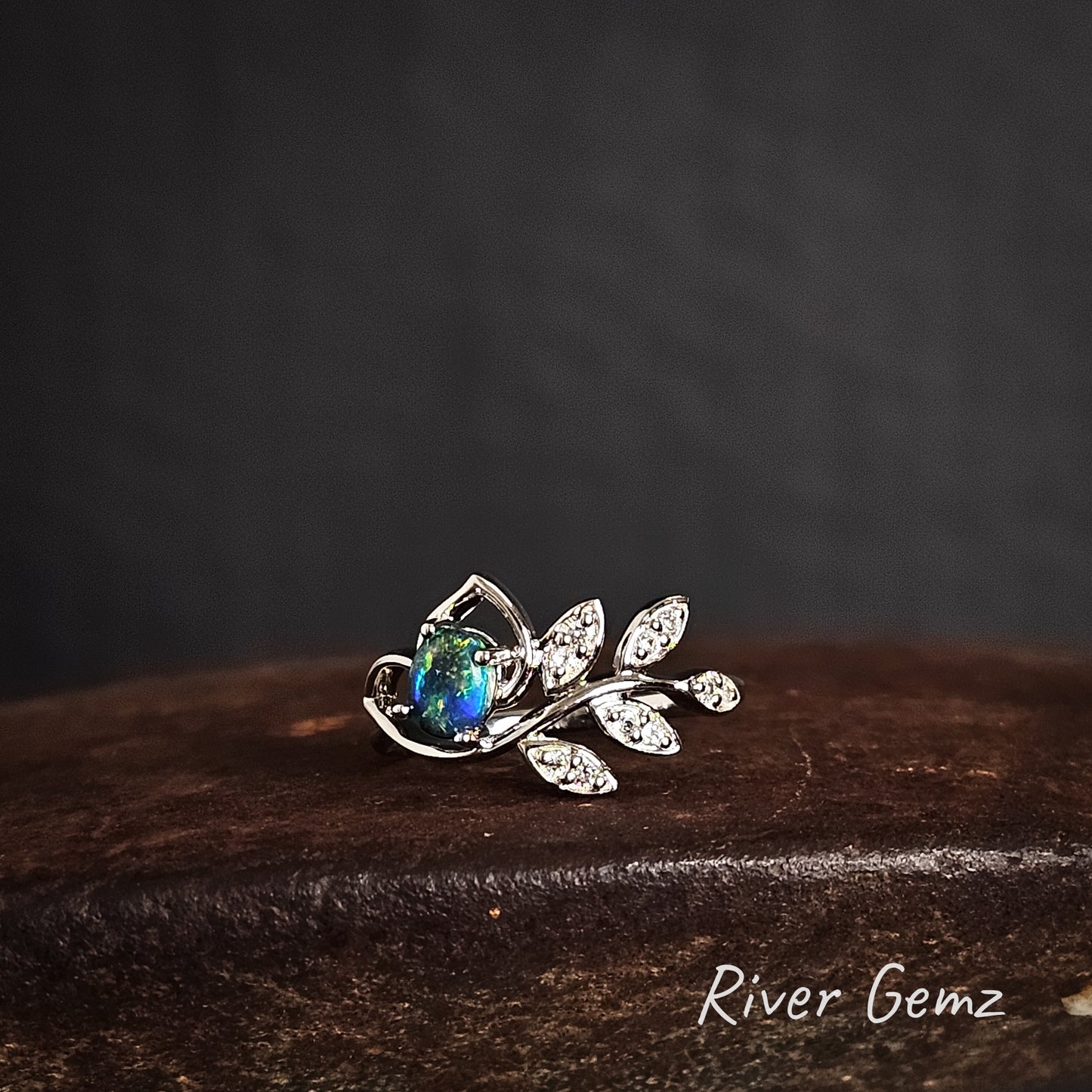 Silver leaf-shaped ring with a green oval shaped black opal on a dark surface, branded 'River Gemz'.