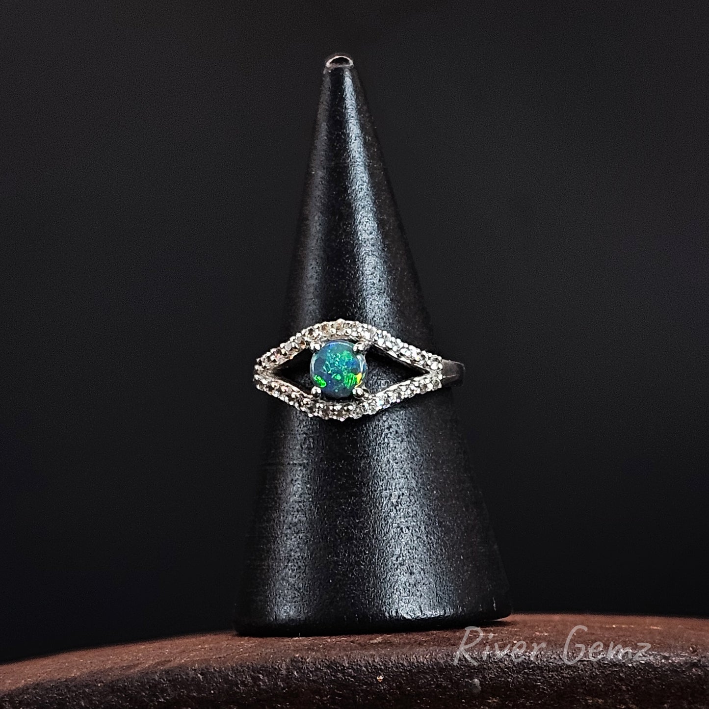 A silver-toned eye-shaped ring with a vibrant green-blue iridescent opal center stone and small clear stones, displayed on a black cone stand against a dark background.