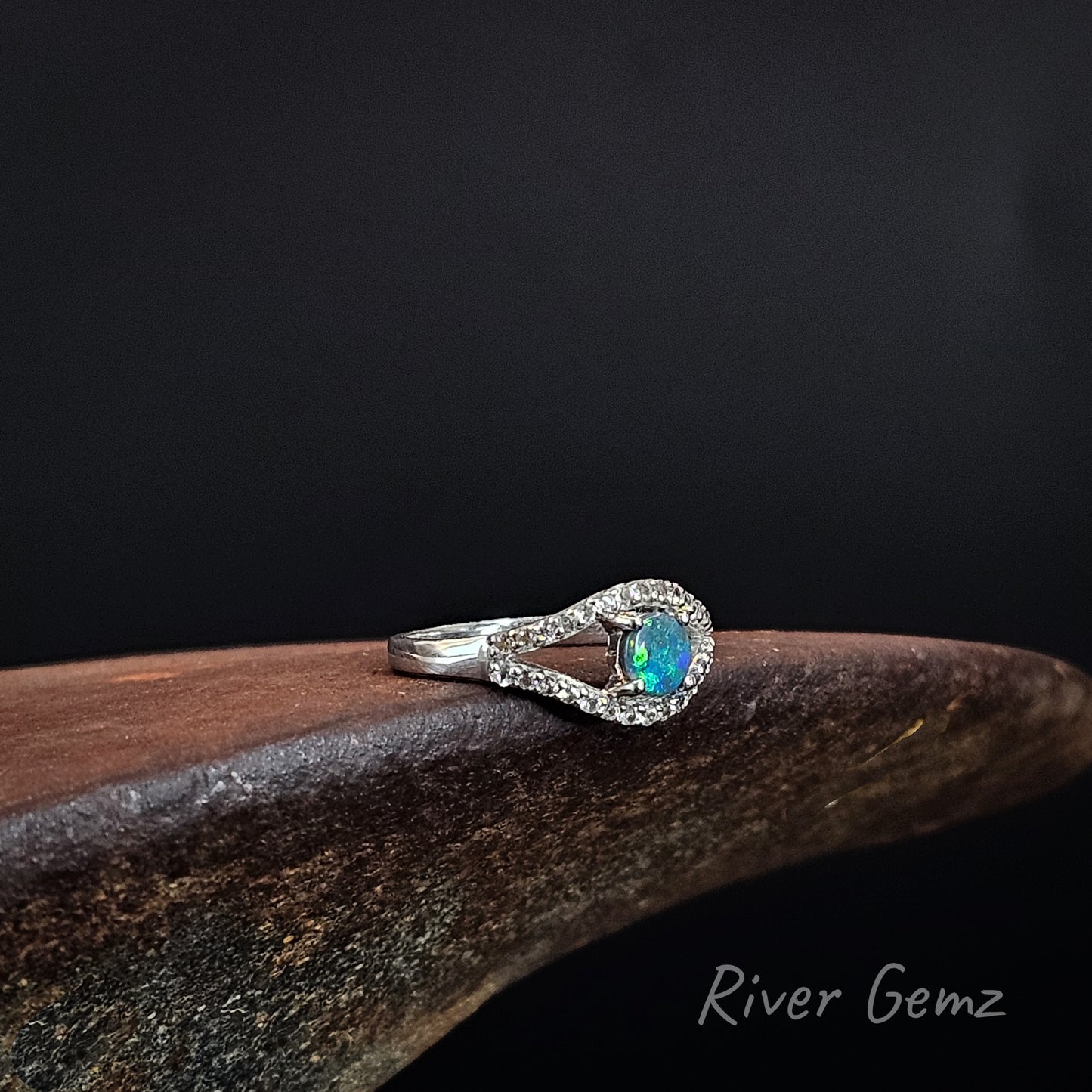 A silver ring featuring a central iridescent blue-green opal surrounded by a halo of small clear stones, resting on a dark brown wooden surface against a black background. The brand name 'River Gemz' is visible in the bottom right corner.