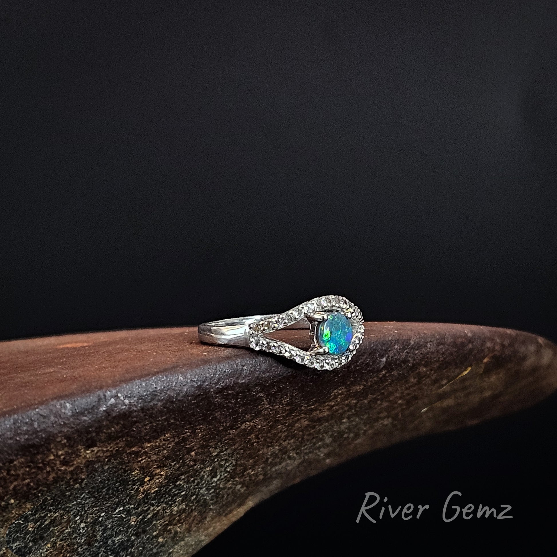 A silver ring featuring a central iridescent blue-green opal surrounded by a halo of small clear stones, resting on a dark brown wooden surface against a black background. The brand name 'River Gemz' is visible in the bottom right corner.