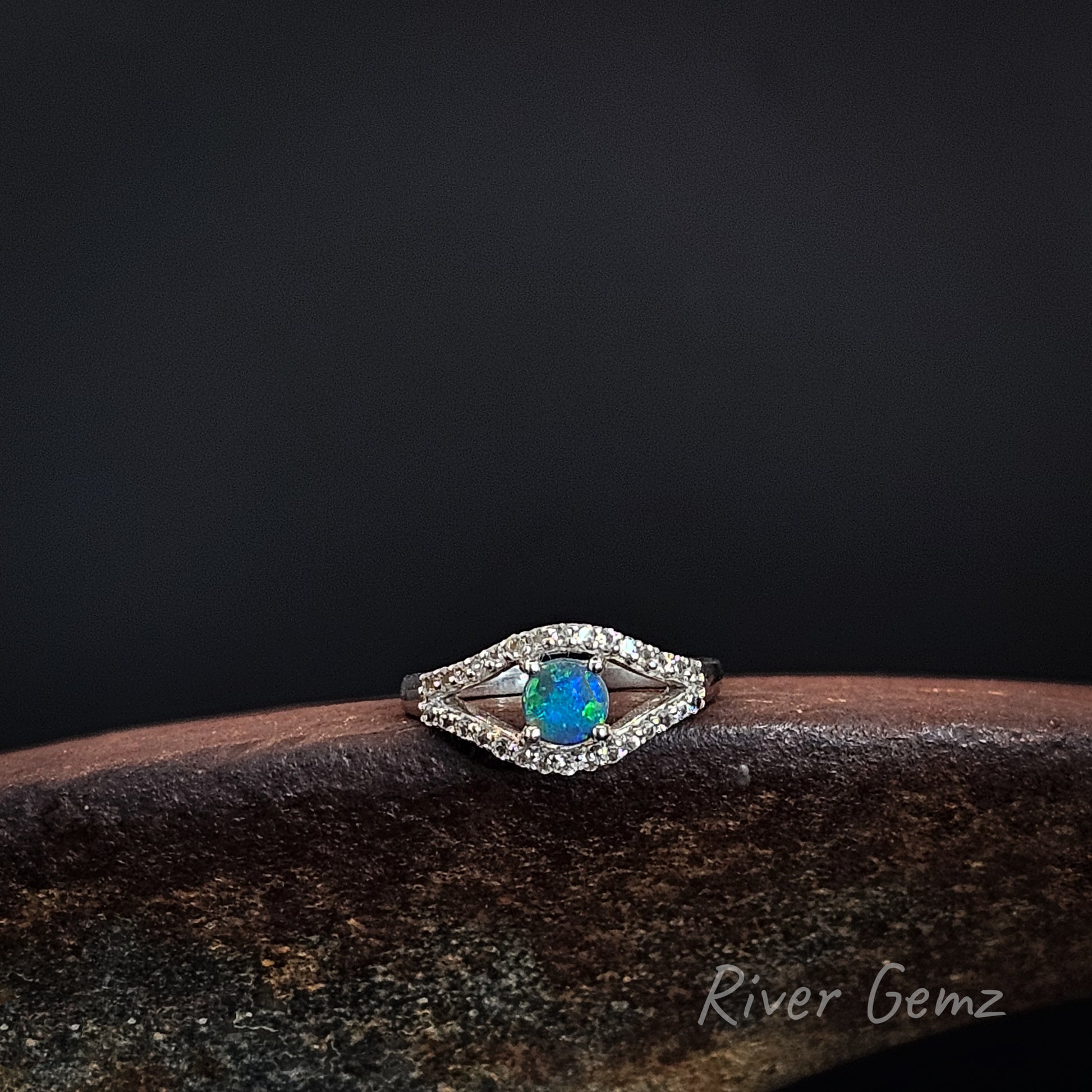 A silver-toned double-halo ring featuring a vibrant blue opal center stone, resting on a dark textured surface against a black background. The brand name 'River Gemz' is visible in the bottom right corner.
