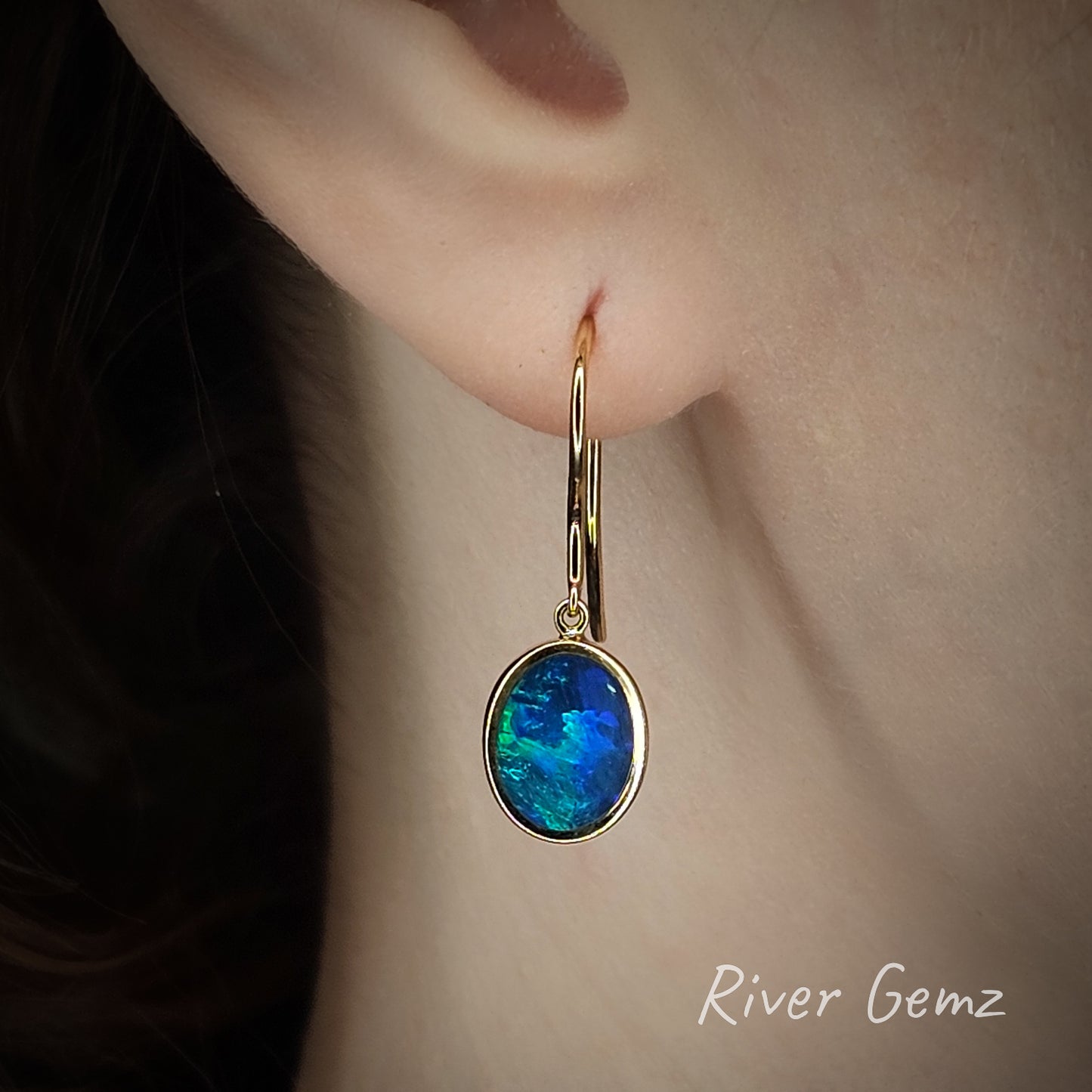Gold hook earring with a blue and green black opal worn by a person, branded 'River Gemz'.