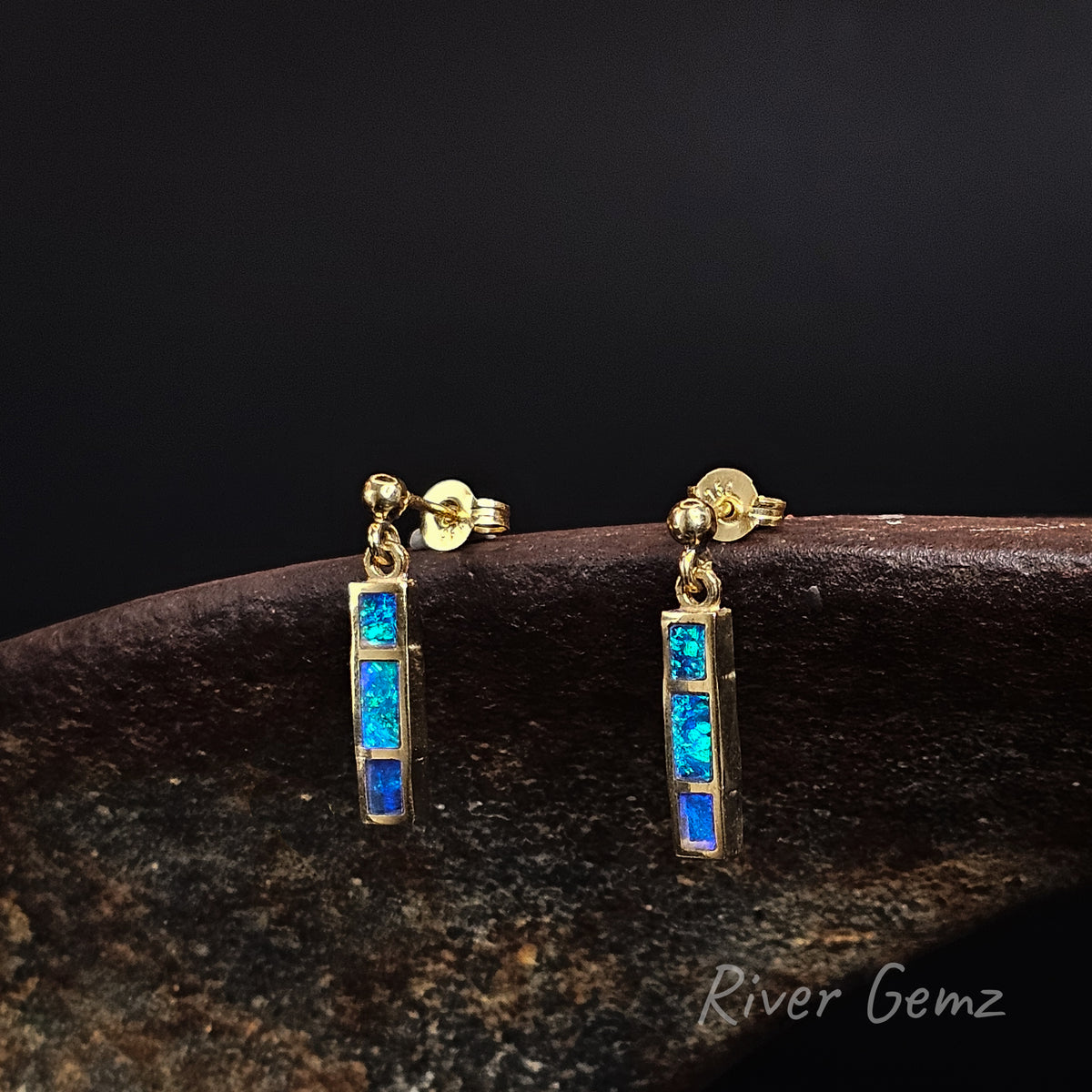 Three rectangular shaped blue and green opals channel set in stud earrings that dangle against a black back ground with 'River Gemz' watermark in the bottom right hand corner.