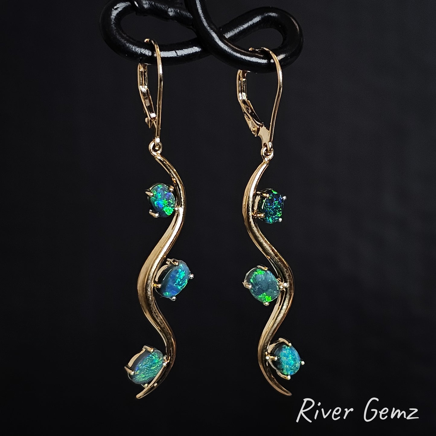 Yellow gold hook earrings with blue and green dark opals on a black background, branded 'River Gemz'.