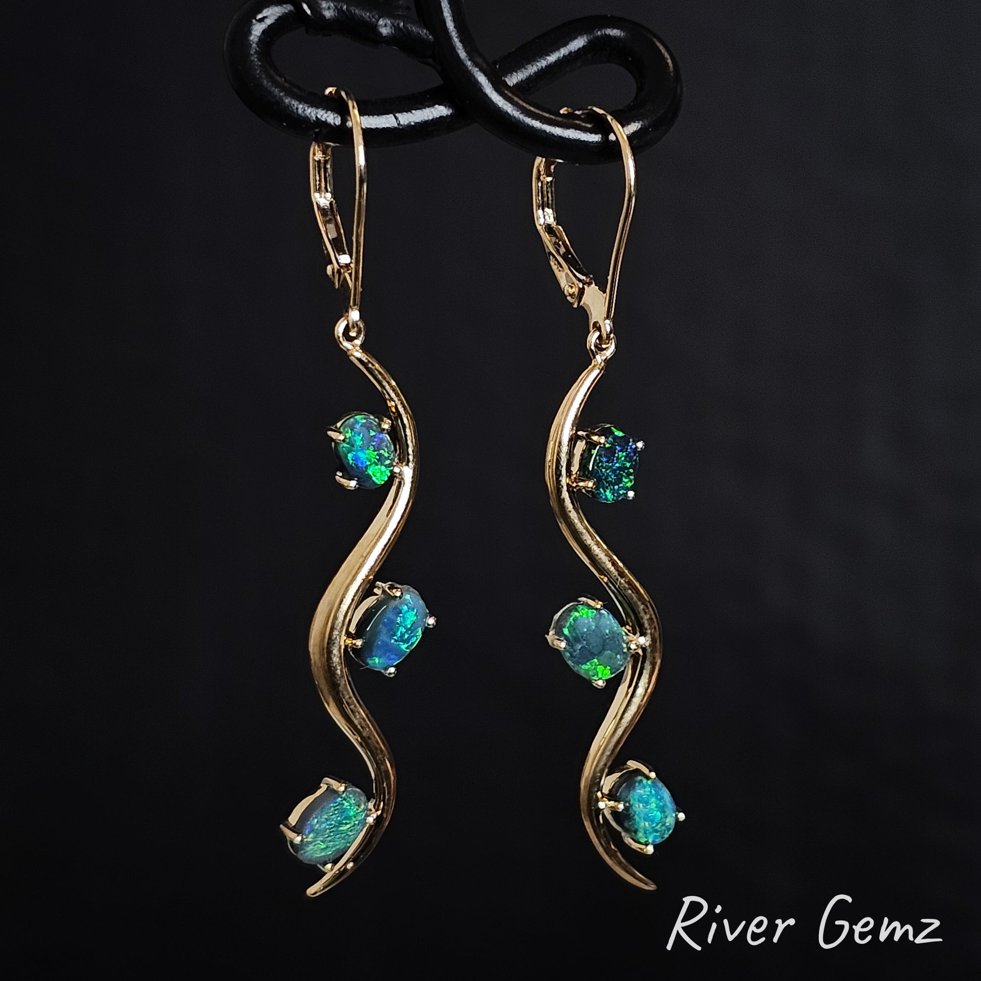 Yellow gold hook earrings with blue and green dark opals on a black background, branded 'River Gemz'.